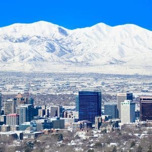 A city skyline on a clear day, with a massive snow-covered mountain range towering in the background.
