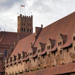Un imponente castello in mattoni rossi con tetti in tegole e una torre dove sventola la bandiera polacca sotto un cielo nuvoloso.