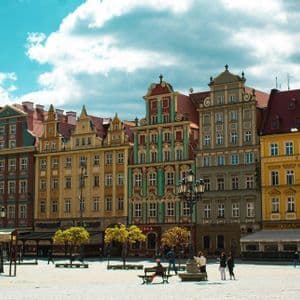 Una fila di edifici colorati e tradizionali in un'ampia piazza acciottolata dove le persone passeggiano e si rilassano sotto un cielo parzialmente nuvoloso.