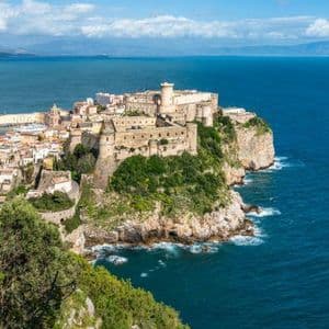 Una storica città fortificata arroccata su una verdeggiante e rocciosa penisola a picco su un vasto mare blu sotto un cielo parzialmente nuvoloso.