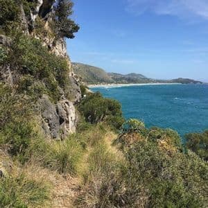 Una vista da un sentiero roccioso a picco sul mare turchese, con costa e colline in lontananza sotto un cielo azzurro.