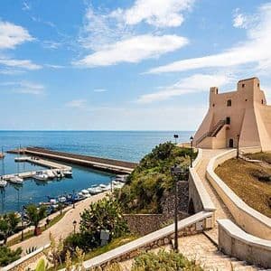 Una fortezza costiera sorge su una collina affacciata su un porto turistico pieno di barche, sotto un cielo parzialmente nuvoloso.