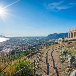 Un sentiero in cima a una collina con antiche rovine si affaccia su una città costiera e sul mare sotto un cielo limpido e soleggiato.