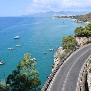 Una strada costiera tortuosa costeggia una scogliera che si affaccia su una baia turchese con diverse imbarcazioni, con un litorale distante sullo sfondo.