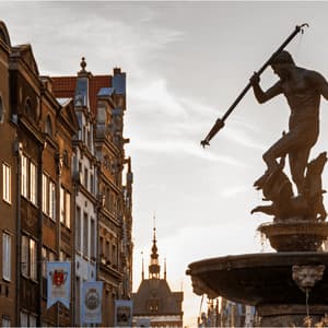 Una grande fontana in bronzo raffigurante una statua di Nettuno con un tridente, sullo sfondo di edifici storici e un cielo al tramonto.