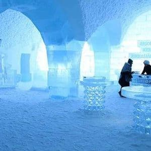 L'intérieur d'un bar entièrement de glace, avec deux personnes en vestes d'hiver se tenant au comptoir de glace, près de tables et d'arches.