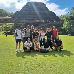 A WeRoad group trip poses for a photo on a grassy lawn with a large, ancient stone pyramid in the background.