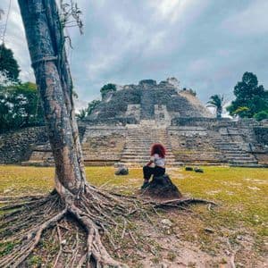 A person with red hair sits on a rock facing a large stone pyramid in a jungle clearing, with a large tree in the foreground.