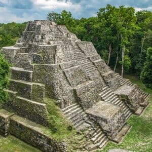 An ancient stone step pyramid rises from a clearing in a dense green jungle under a cloudy sky.