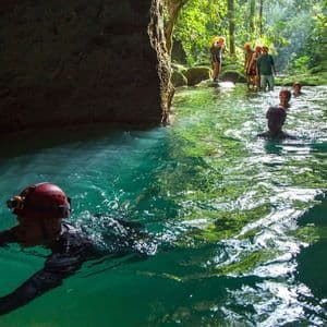 A WeRoad group trip wearing helmets swims through a turquoise river at the entrance of a jungle cave.