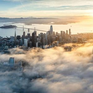 Aerial view of a city skyline with skyscrapers emerging from a dense blanket of fog as the sun rises over the water.