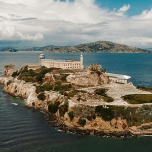 An aerial view of a historic prison complex on a rocky island, surrounded by water with distant hills under a cloudy sky.