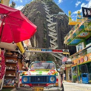 A colorful vintage car parked on a street below a large rock mountain with a prominent zig-zagging staircase leading up its side.
