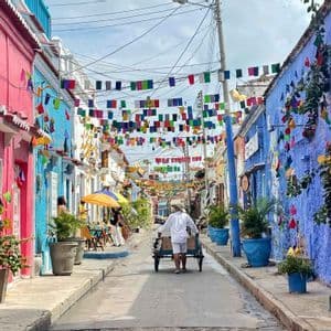 A man pushes a cart down a narrow street lined with colorful buildings and decorated with overhead flags.