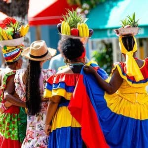 A group of women seen from behind in colorful, traditional dresses, balancing bowls of fresh fruit on their heads.