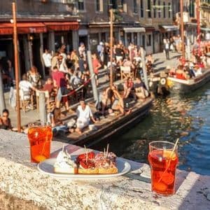 Due cocktail arancioni e stuzzichini su un davanzale in pietra, con vista su un vivace canale dove gente socializza su barche e in un caffè sul lungomare.