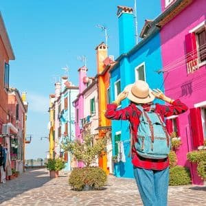 Una persona con uno zaino, vista da dietro, si tiene il cappello mentre è in piedi su una strada fiancheggiata da edifici colorati.