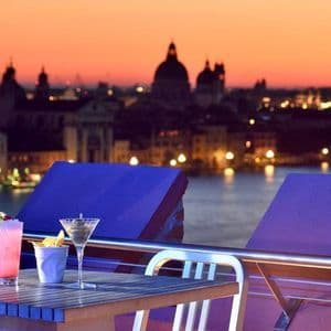 Due cocktail su un tavolo in un bar panoramico con vista sullo skyline illuminato della città e sull'acqua al tramonto.