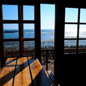 Un tavolo di legno in una stanza buia, posizionato accanto a grandi finestre a riquadri, offre una vista soleggiata su una costa rocciosa e sull'oceano.