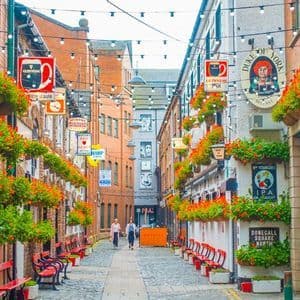 Un vicolo stretto e acciottolato, fiancheggiato da edifici in mattoni, insegne di pub, cesti di fiori appesi e lucine.