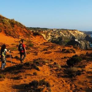 Due escursionisti con zaini percorrono un sentiero costiero sabbioso e arancione, con scogliere a picco sull'oceano.