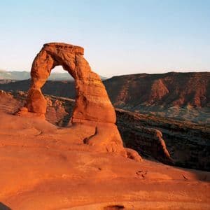 A large, natural sandstone arch glows in the warm light of sunrise over a rocky desert landscape with mountains in the distance.