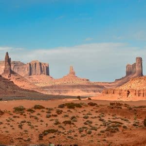 A wide shot of a red sand desert valley with large sandstone buttes and mesas rising in the distance under a blue sky.