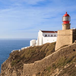 Un faro di pietra con la sommità rossa e edifici bianchi adiacenti si ergono su una scogliera rocciosa a picco sull'oceano sotto un cielo azzurro.