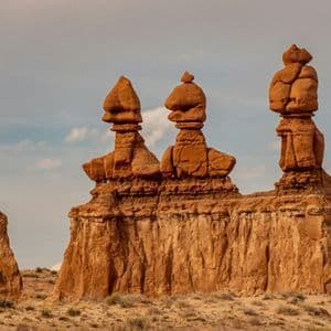 Three large, reddish-brown hoodoo rock formations stand in a desert landscape under a partly cloudy sky.