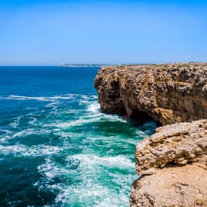Scogliere aspre color ocra dominano un mare turchese e azzurro impetuoso con onde bianche che si infrangono sotto un cielo sereno.
