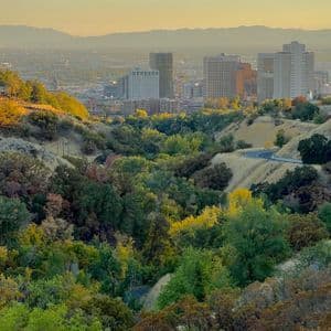 A city skyline with skyscrapers and a capitol building viewed from rolling hills with colorful autumn trees at sunset.