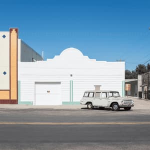 A vintage white station wagon parked on a street in front of minimalist storefronts under a clear blue sky.