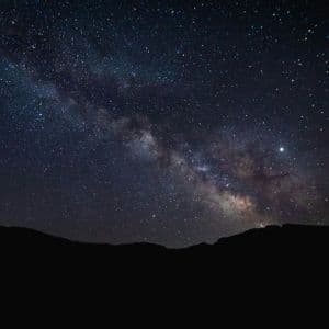The Milky Way galaxy arcs across a dark, star-filled night sky above the silhouette of mountains.