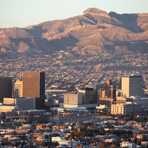 An expansive city skyline with downtown buildings is nestled at the base of a large, arid mountain range at golden hour.