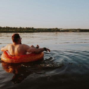 A shirtless man seen from behind relaxes in an orange inflatable ring on calm water at sunset.