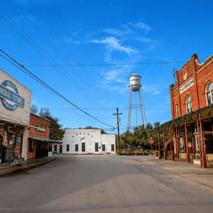 An empty street in a historic town features old-fashioned storefronts and a tall water tower under a clear blue sky.