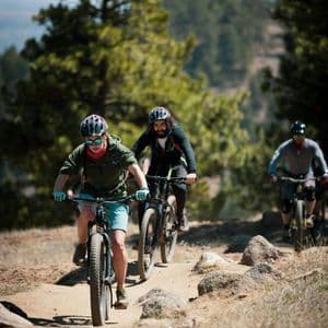 A WeRoad group trip of people riding mountain bikes on a dirt trail through a sunny, forested area.