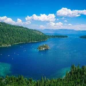 A high-angle view of a deep blue lake with boats surrounding a small, tree-covered island, all enclosed by forested hills under a blue sky.
