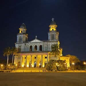 Eine große, historische Kathedrale ist nachts beleuchtet, davor ein weiter, leerer Kopfsteinpflasterplatz, vor einem dunklen Himmel mit hellem Mond.