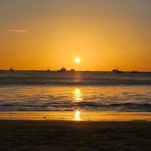 Die Sonne versinkt über dem Meer, Boote zeichnen sich als Silhouetten am Horizont ab, während sich ihr Schein auf dem nassen Sand spiegelt.