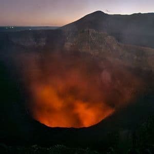 Weitblick auf einen Vulkankrater bei Sonnenuntergang, mit glühender orangefarbener Lava und aufsteigendem Rauch.
