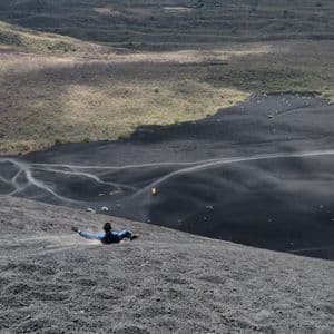 Eine Person rutscht einen steilen Hang aus schwarzem Vulkangestein hinab, mit Blick auf ein großes Tal darunter.