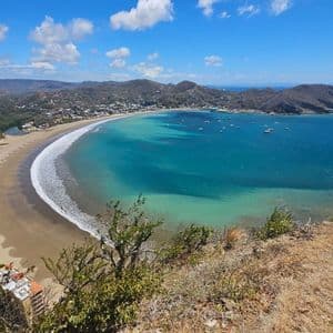 Vogelperspektive auf eine Küstenstadt an einem geschwungenen Sandstrand, angrenzend an eine türkisfarbene Bucht mit Booten, unter einem teils bewölkten Himmel.