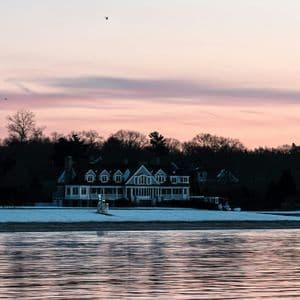 A large white house sits on a snowy shoreline across from a body of water, under a pink and purple sunset sky.