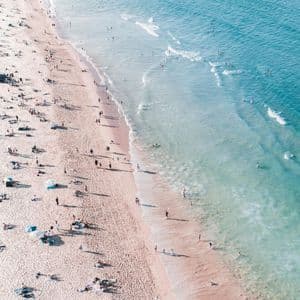 An aerial view of many people relaxing on a sandy beach and swimming in the turquoise ocean.