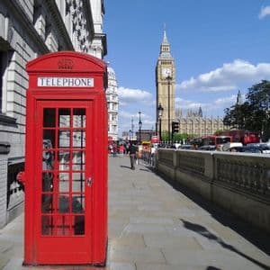 A red telephone booth on a sidewalk with the Big Ben clock tower in the background on a sunny day.