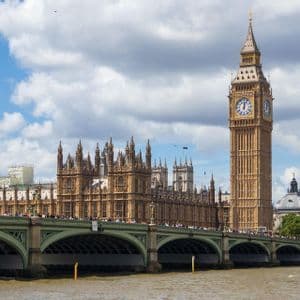 A large clock tower and a historic building stand behind a crowded stone bridge over a river under a partly cloudy sky.