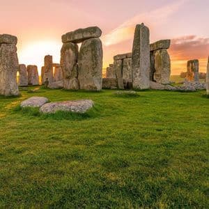 The sun sets behind a circle of ancient standing stones on a lush green field.