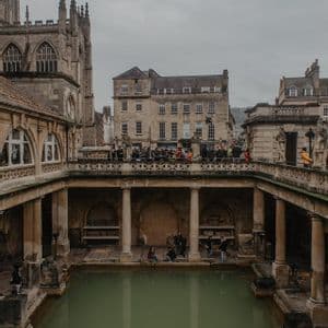 People stand on a terrace overlooking a large thermal pool surrounded by historic stone architecture under a cloudy sky.
