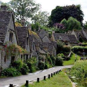 A row of historic stone cottages with slate roofs lines a narrow road next to a small stream in a lush, green village.